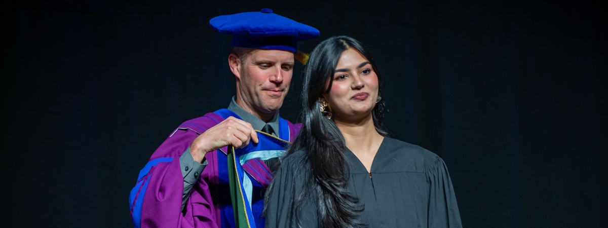 A professor in purple and blue convocation gown putting a ribbon on a graduate in black