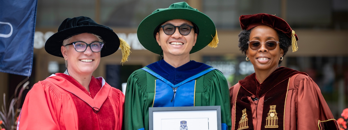A graduate in green convocation gown holding his degree while 2 professors in red and brown gown stand on either side of him